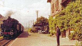 <p>Der Bahnhof in Distelhausen 1957 mit einfahrender Dampflok und Bahnagent August Müller. Die Blumenpracht rund um das Bahnhofsgebäude brachte dem Standort zahlreiche Auszeichnungen ein.</p>