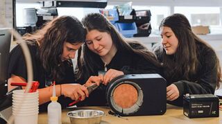 Selina Volkert, Ivana und Ivona Petkovska von der Eduard-Mörike-Gemeinschaftsschule in Bad Mergentheim arbeiten am leuchtenden und wasserwärmenden Hundenapf. Unterstützt werden sie dabei von ecom-Mitarbeiter Maik Gutte.