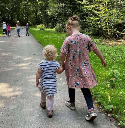 Zwei Mädchen gehen einen Waldweg entlang, laufen auf eine Gruppe Kinder zu. Das größere, selbstsichere Kind hat das kleine an die Hand genommen, das ihm vertrauensvoll folgt. „Komm mit“, scheint das ältere Mädchen zu sagen. „Wir gehören doch alle zusammen!“ Inklusion ist: Wenn Behinderung keine Rolle spielt.
