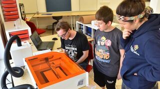Werkstattleiterin Sophie Krimmer schaut zusammen mit den beiden Jugendlichen Marlon (stehend) und Florian (sitzend), ob es gelingt mithilfe des Lasercutters ein Modellauto aus Pappe herzustellen.