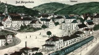 Die Postkarte zeigt den Bahnhof mit dem Uhrtürmchen auf dem Mittelbau um 1900 zusammen mit den beiden Dienstgebäuden der Bahnbeamten und Arbeiter gleich rechts daneben, die 2012 abgebrochen wurden. Gegenüber dem Bahnhof befand sich eine kleine Grünanlage und dahinter das Krankenhaus Carolinum.