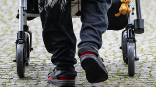 Ein Mann ist mit seinem Rollator auf einem gepflasterten Weg unterwegs.