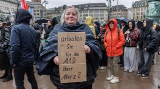Eine Demonstrantin in Hamburg stellt auf einem Pappschild eine Frage an Bundeskanzler Friedrich Merz (CDU).